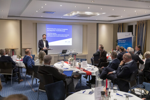 Wirtschaftsminister Claus Ruhe Madsen steht vor einer Leinwand in einem Raum mit geladenen Gästen und hält einen Vortrag.